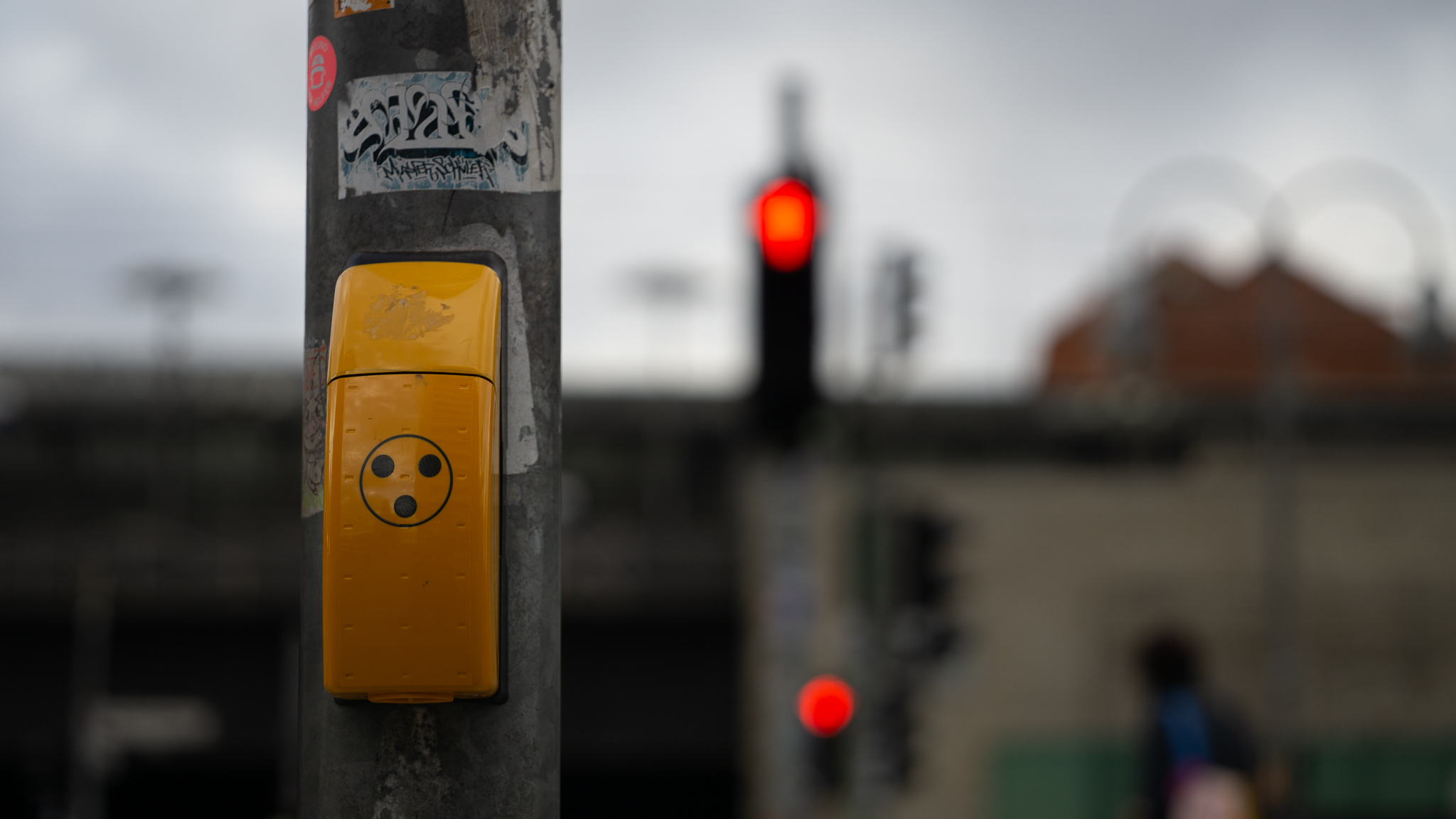 A yellow pedestrian crossing button on a grey pole, with a blurred red traffic light behind it